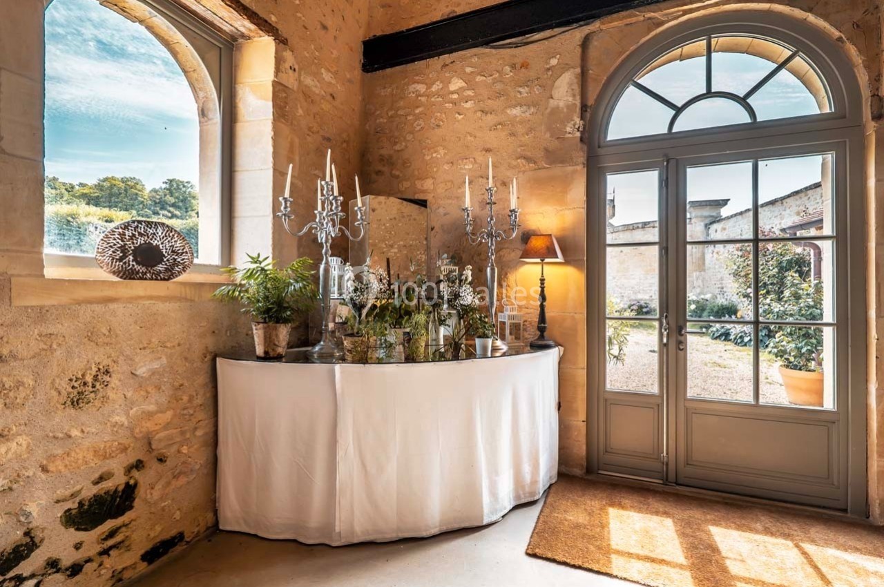Console décorative avec plantes, chandeliers et lampe, placée près d'une fenêtre et d'une porte dans une pièce en pierre.