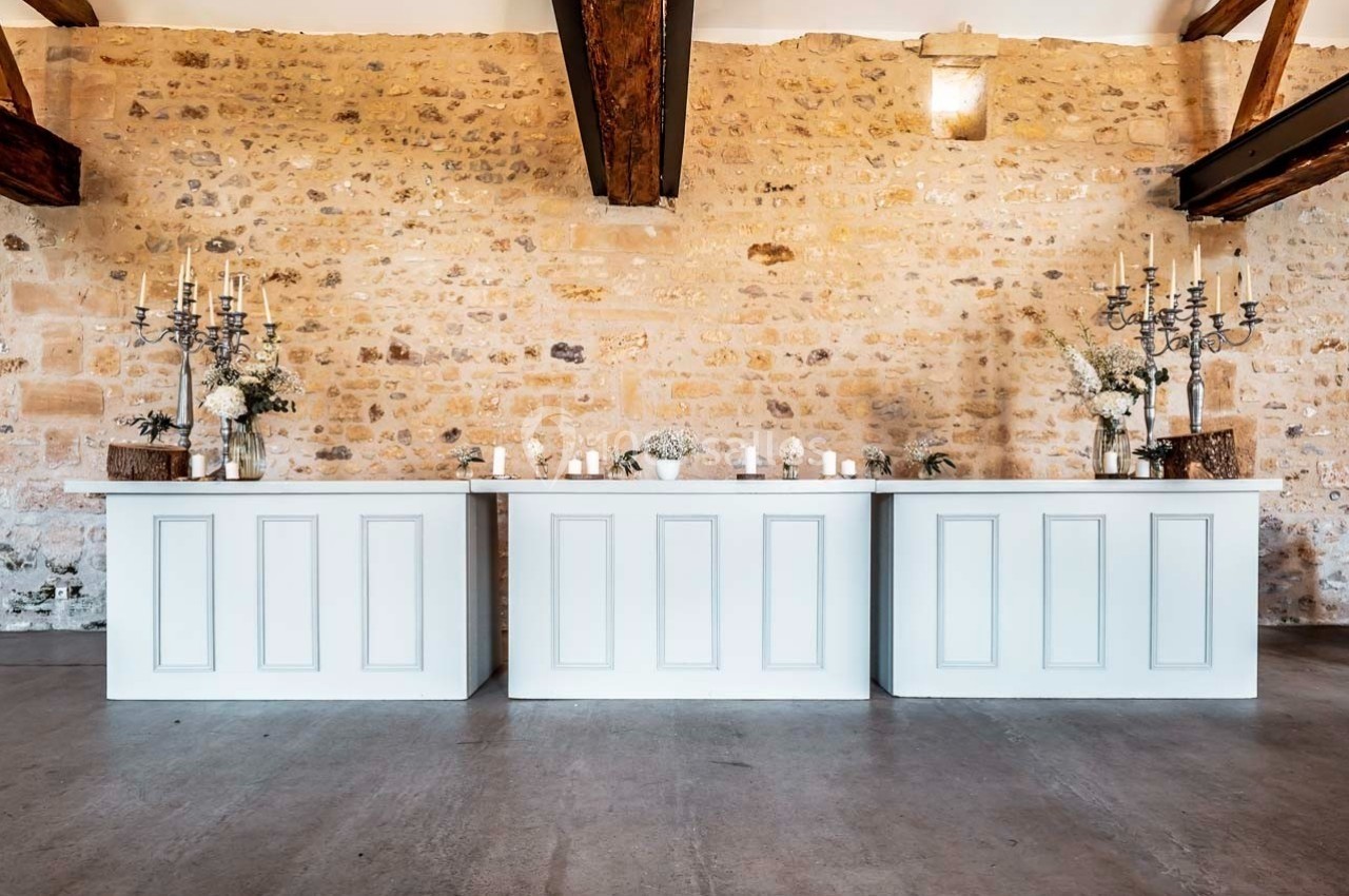 Trois comptoirs blancs décorés de bougies, chandeliers et fleurs, devant un mur en pierre dans une salle lumineuse.