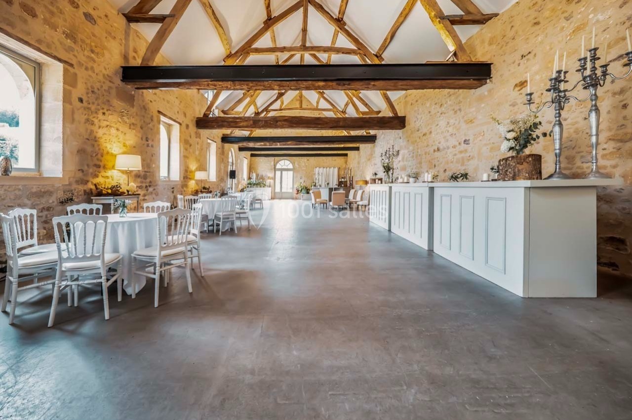 Salle lumineuse avec murs en pierre, poutres apparentes, tables dressées et comptoir blanc dans un cadre élégant.