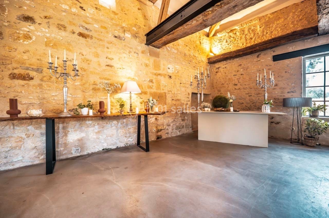 Salle en pierre avec poutres apparentes, tables décorées de chandeliers et plantes, éclairée par une fenêtre.