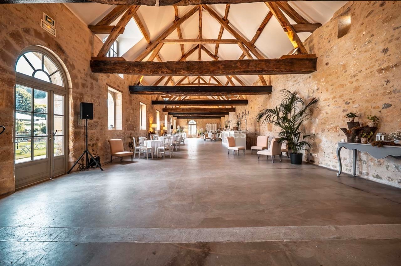 Salle spacieuse avec poutres apparentes, murs en pierre, grandes fenêtres et mobilier disposé pour un événement.