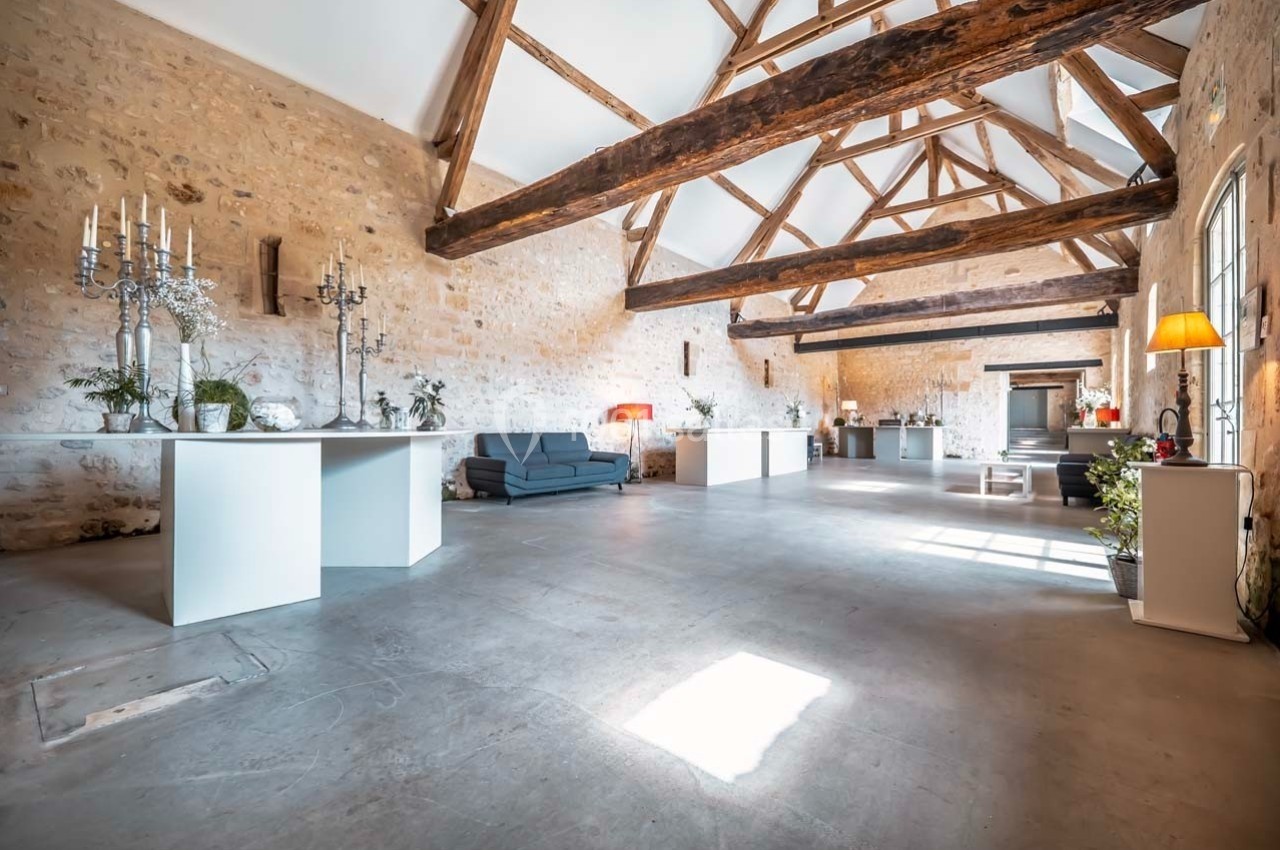 Salle spacieuse avec murs en pierre, poutres apparentes, mobilier moderne et lumière naturelle abondante.