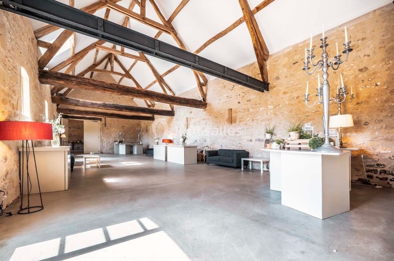 Salle lumineuse avec poutres apparentes, murs en pierre, mobilier moderne et grandes fenêtres laissant entrer la lumière.