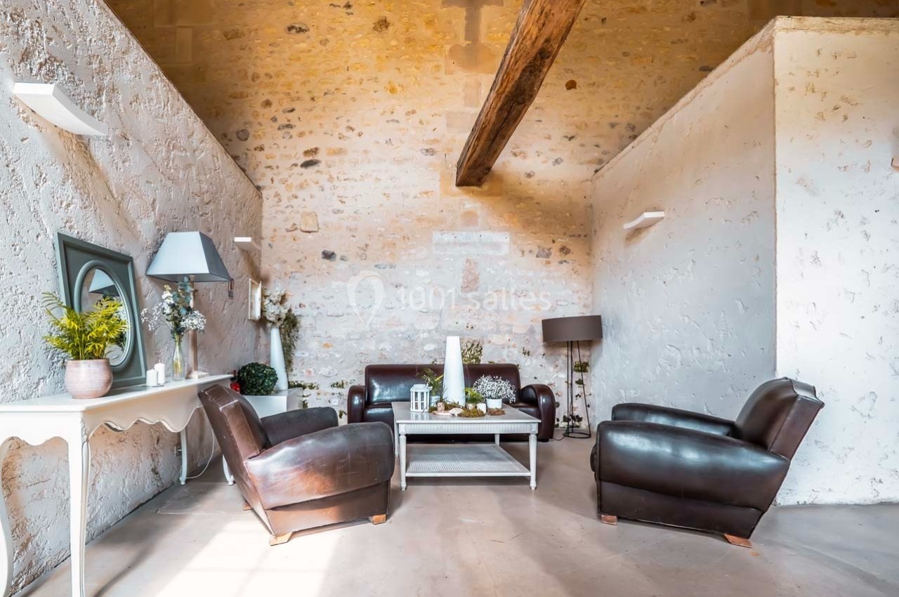 Salon lumineux avec murs en pierre, poutre apparente, mobilier en cuir marron et table basse décorée.