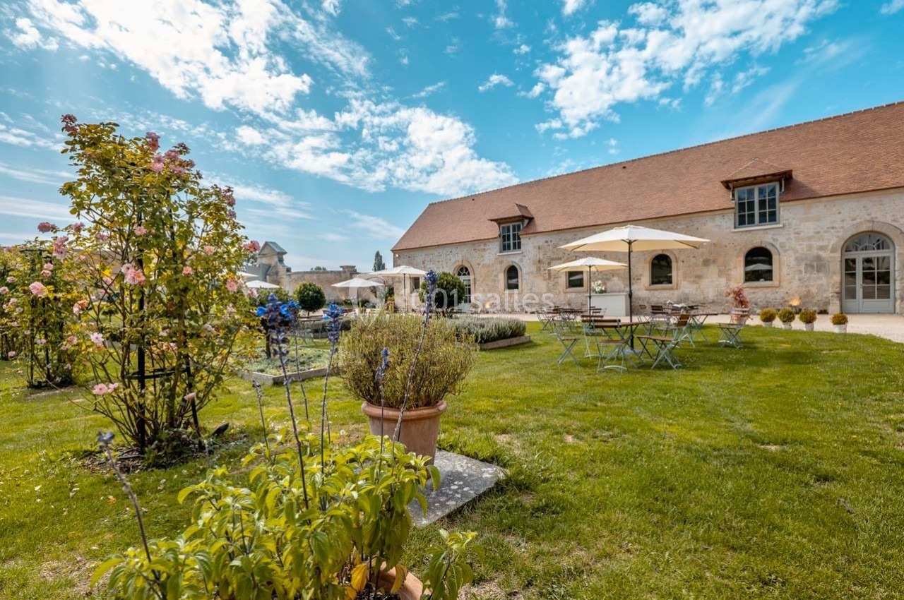 Jardin avec tables et parasols devant un bâtiment en pierre sous un ciel bleu partiellement nuageux.