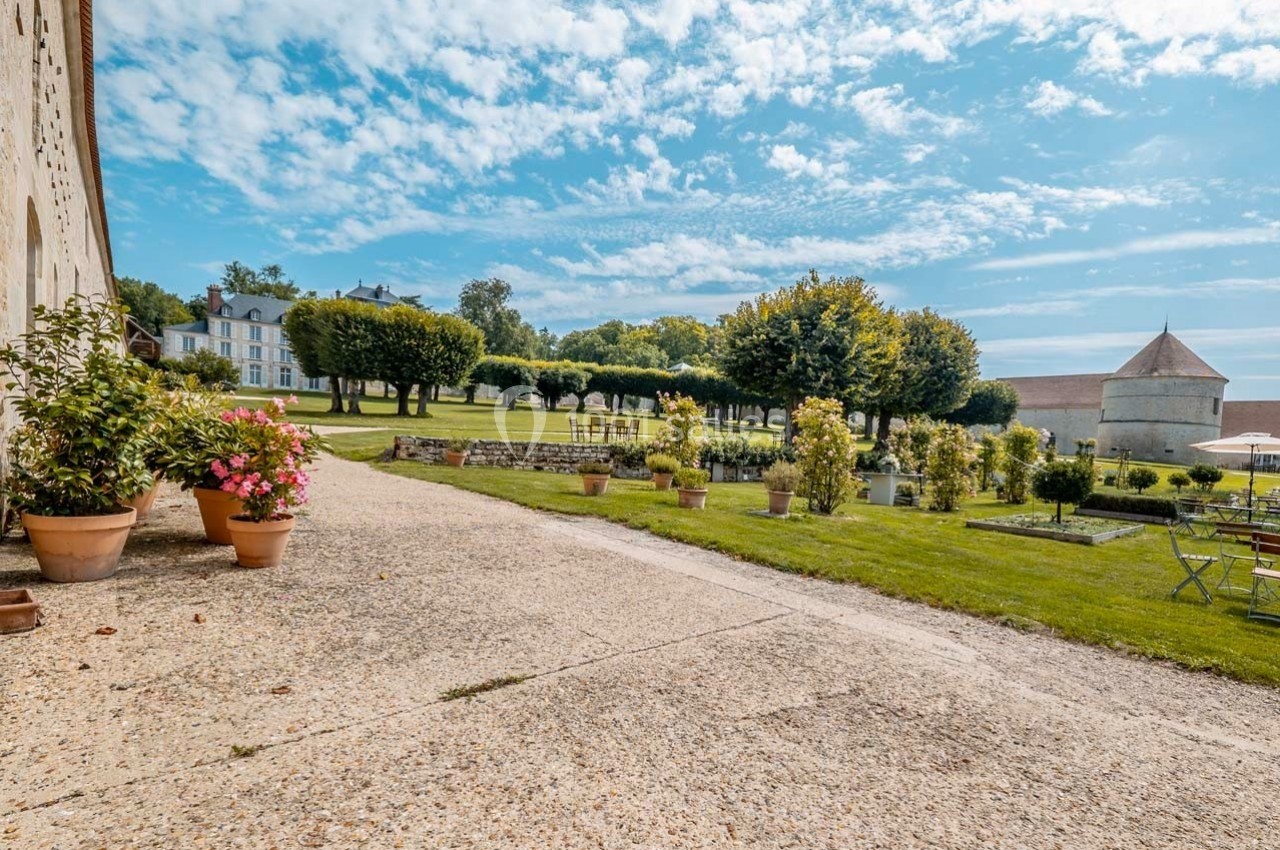 Allée pavée bordée de pots de fleurs menant à un jardin avec pelouse, arbres et bâtiments en arrière-plan.