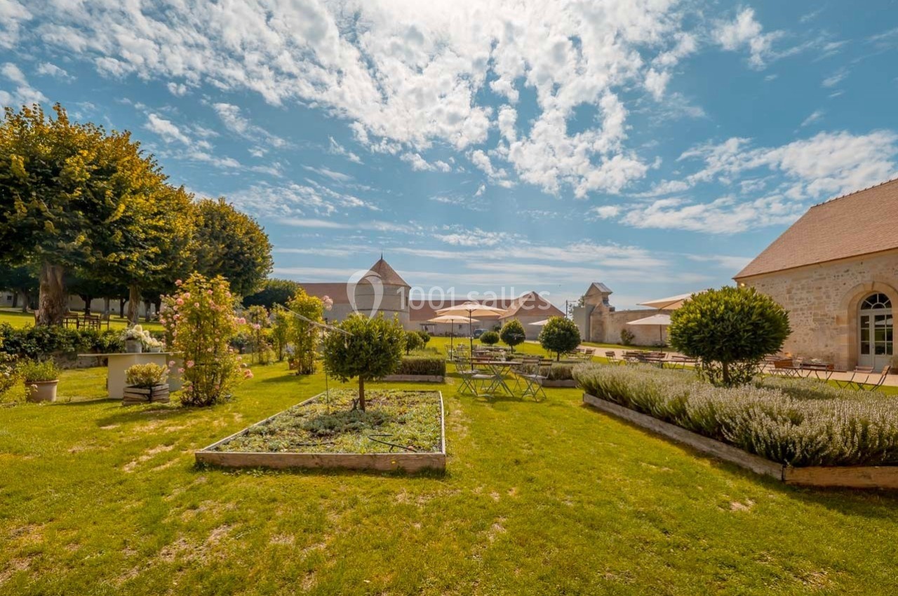 Jardin aménagé avec parterres de plantes, tables et chaises, entouré de bâtiments en pierre sous un ciel partiellement…