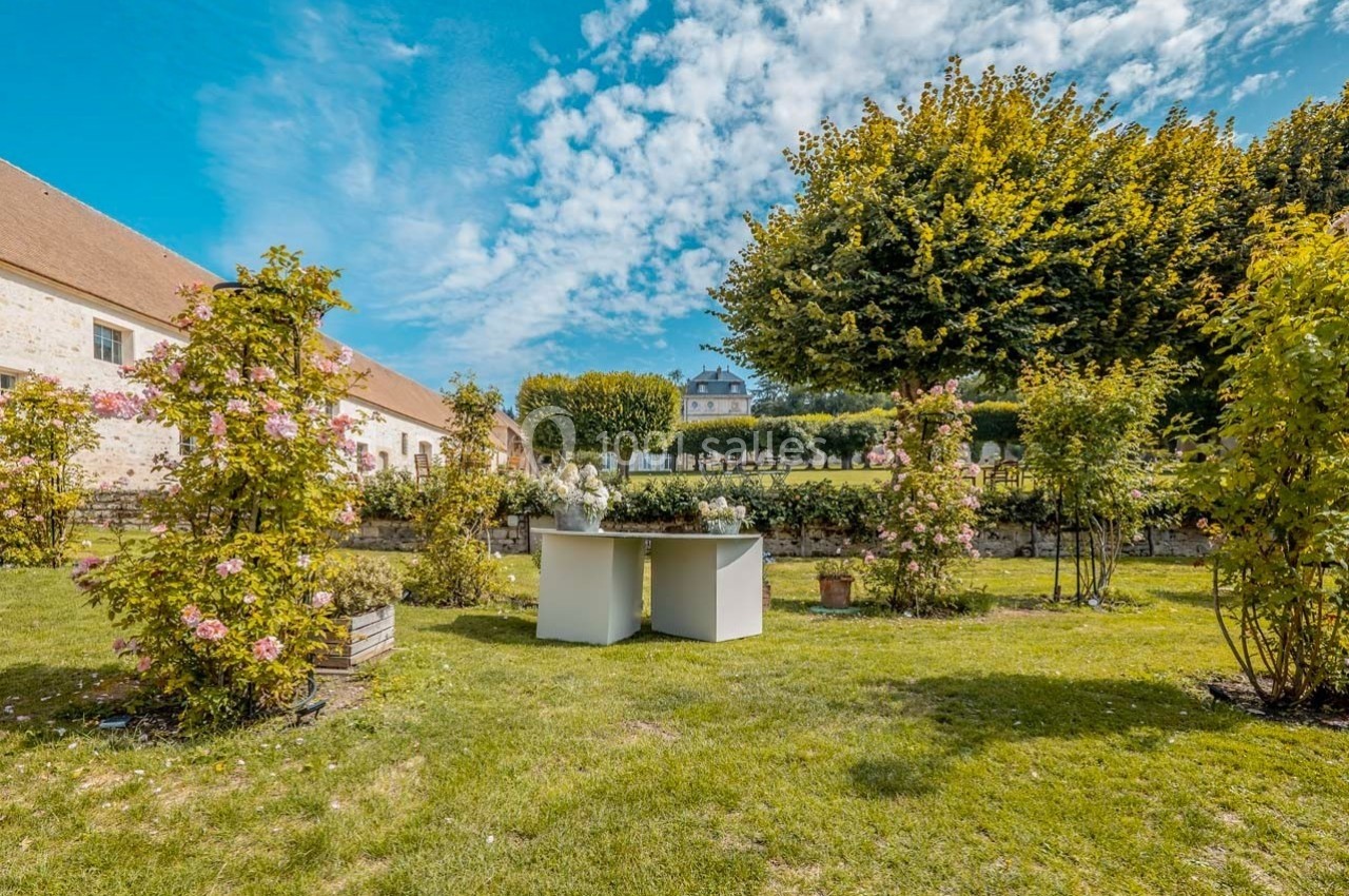Jardin verdoyant avec une table blanche entourée de rosiers, devant un bâtiment en pierre et des arbres.