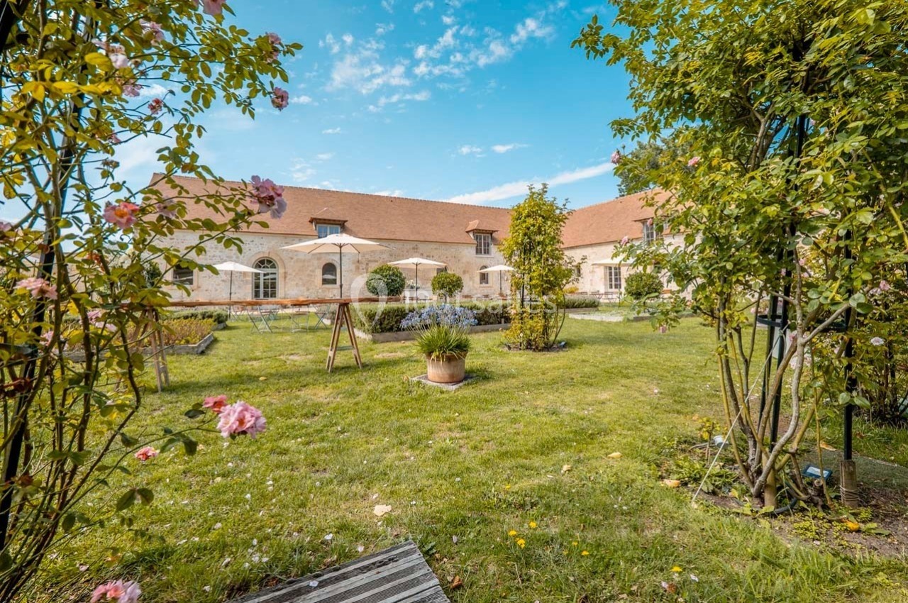 Jardin verdoyant avec fleurs, arbres et mobilier de jardin devant un bâtiment en pierre sous un ciel bleu.