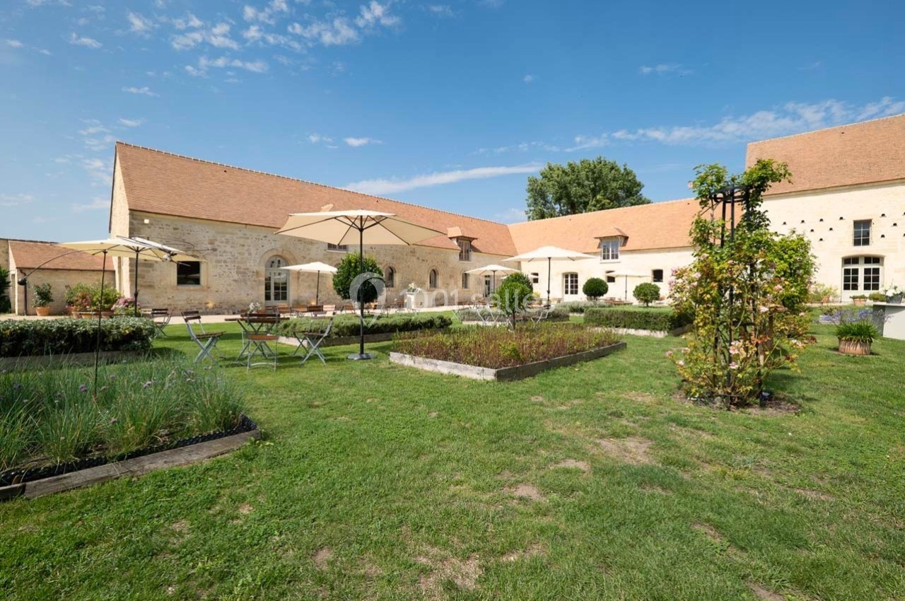Cour intérieure d'un bâtiment en pierre avec jardin, parasols, tables et chaises sous un ciel bleu dégagé.