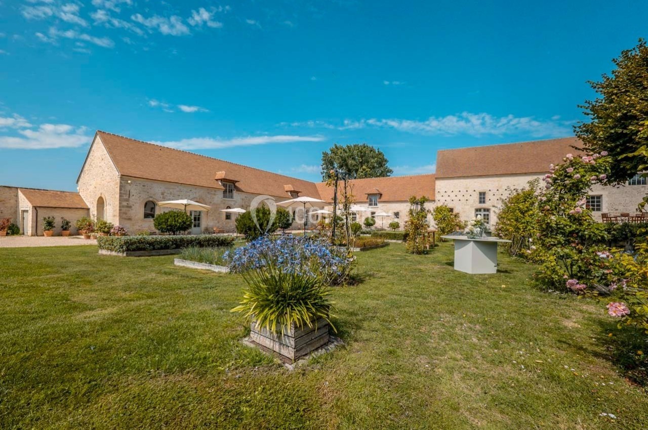 Cour d'une grande bâtisse en pierre avec jardin fleuri, pelouse bien entretenue et ciel dégagé en arrière-plan.