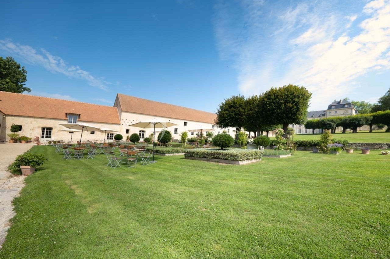Cour verdoyante avec tables et chaises en métal, entourée de bâtiments en pierre sous un ciel dégagé.