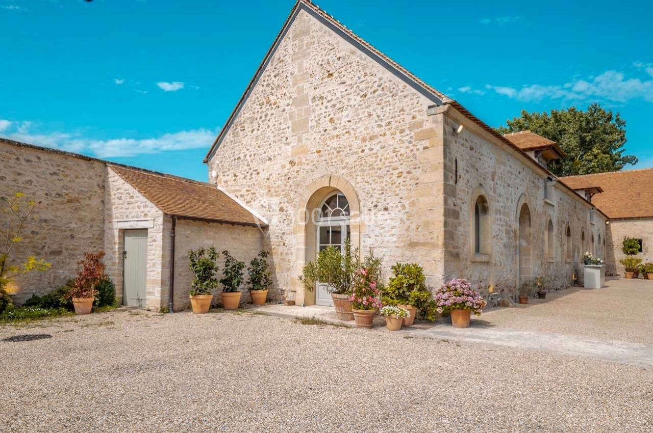 Bâtiment en pierre avec toit en tuiles, entouré de pots de fleurs, dans une cour en gravier sous un ciel bleu.