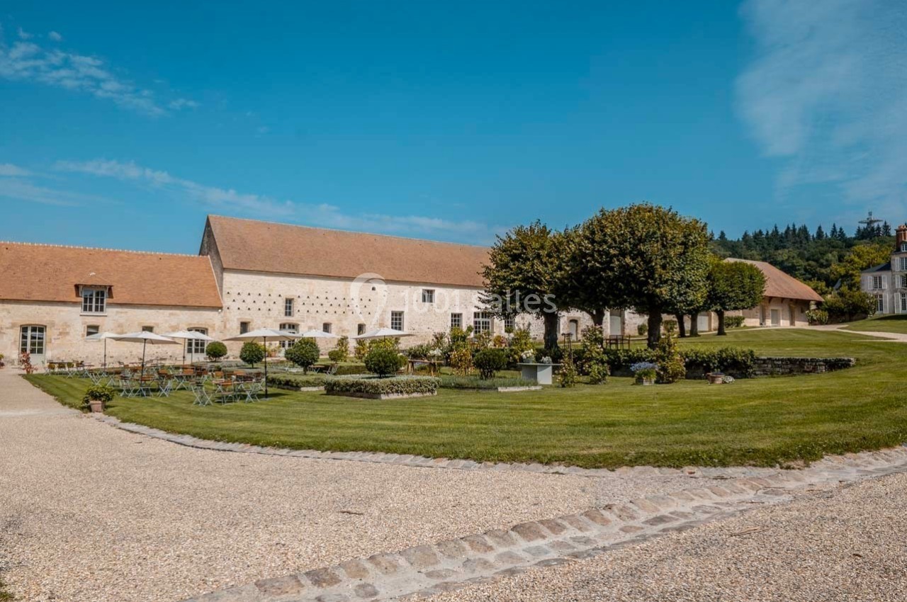 Bâtiment en pierre avec toit en tuiles, entouré d'un jardin aménagé et d'une pelouse sous un ciel bleu.