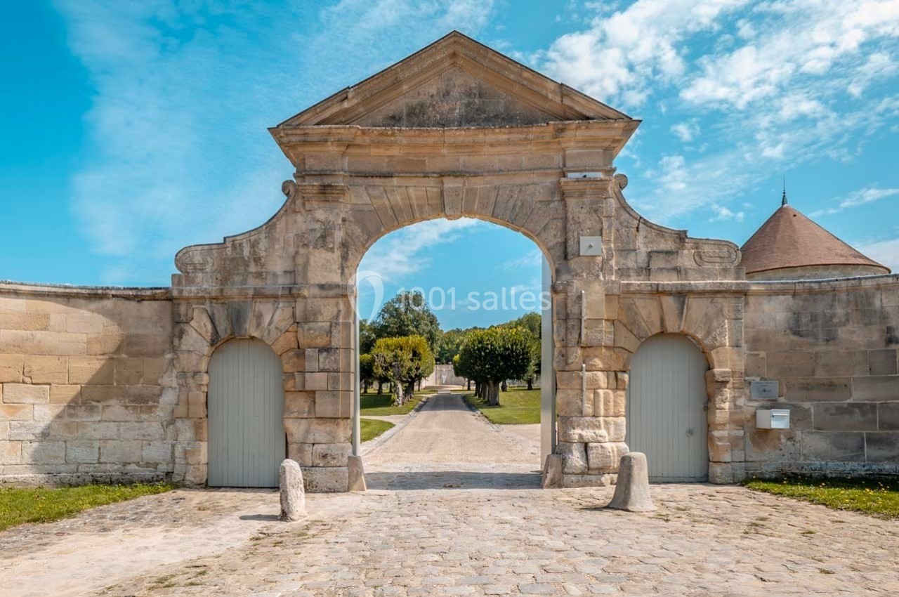 Porte en pierre ancienne avec deux passages, donnant sur une allée bordée d'arbres sous un ciel bleu.