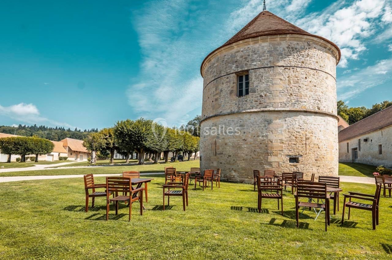 Chaises et tables en bois disposées sur une pelouse devant une tour en pierre sous un ciel bleu.