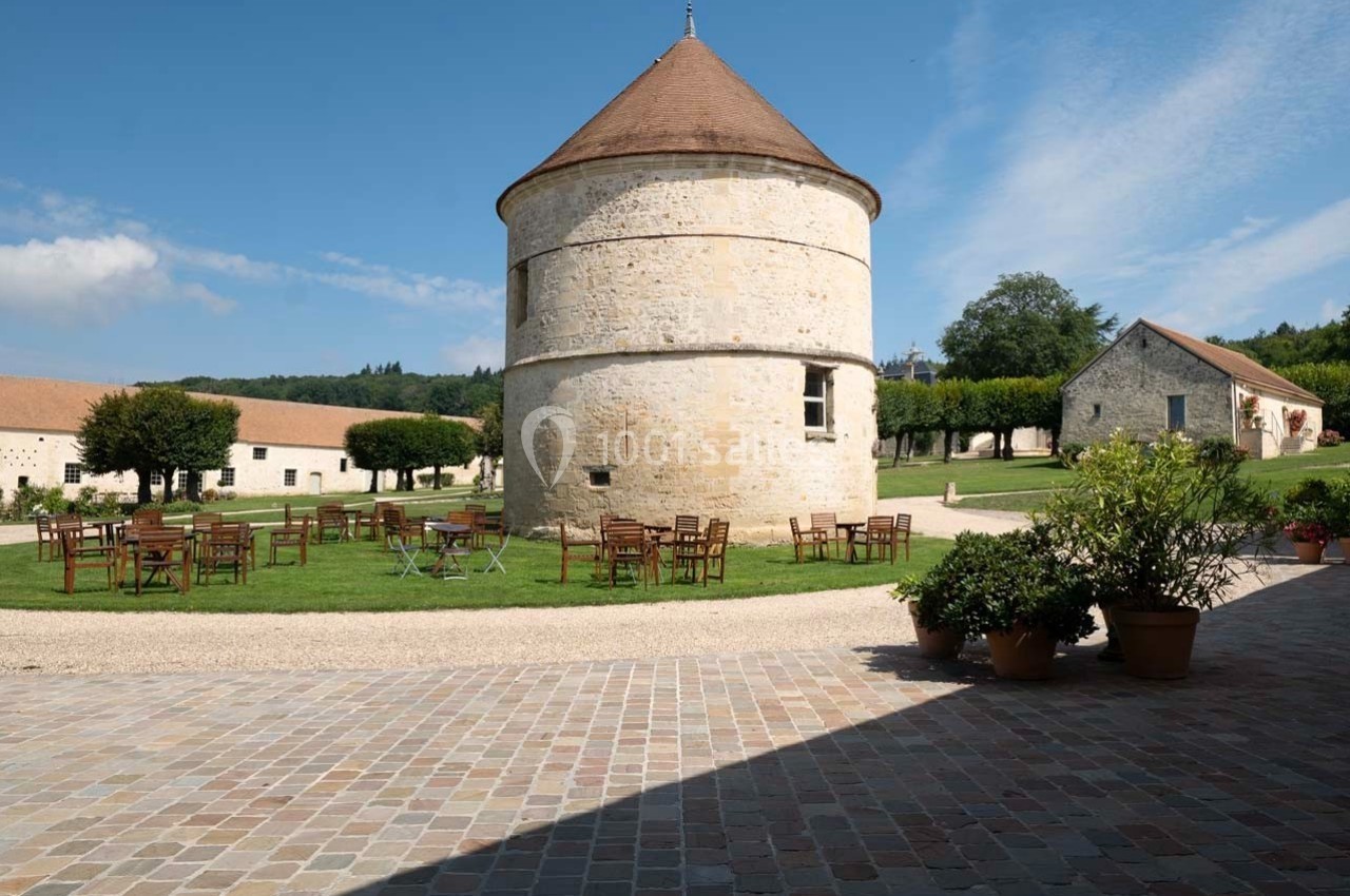 Tour en pierre avec toit conique entourée de tables et chaises en bois, située dans un espace verdoyant et pavé.