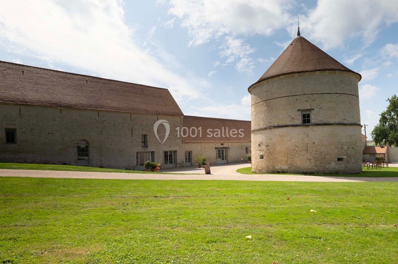 Bâtiments en pierre avec un toit en tuiles, comprenant une tour ronde et une cour herbeuse sous un ciel partiellement…