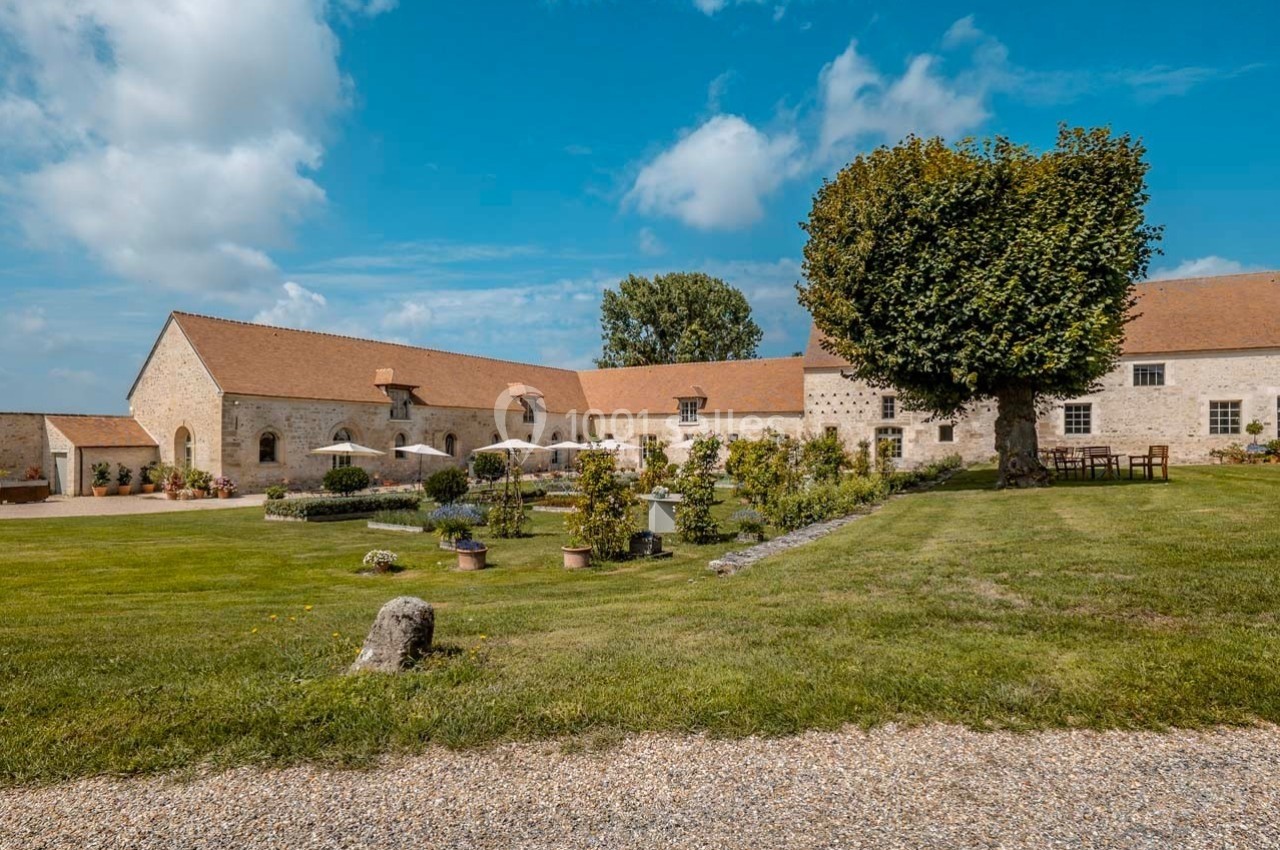 Cour d'une bâtisse en pierre avec toit en tuiles, grand arbre, pelouse, fleurs et mobilier de jardin sous un ciel bleu.