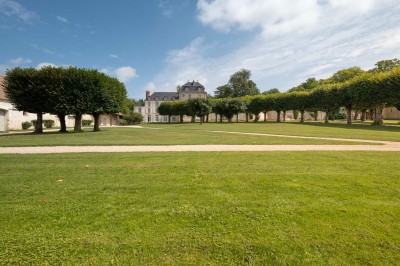 Grand bâtiment classique entouré d'une pelouse bien entretenue, avec un arbre imposant et un petit bassin au premier plan.