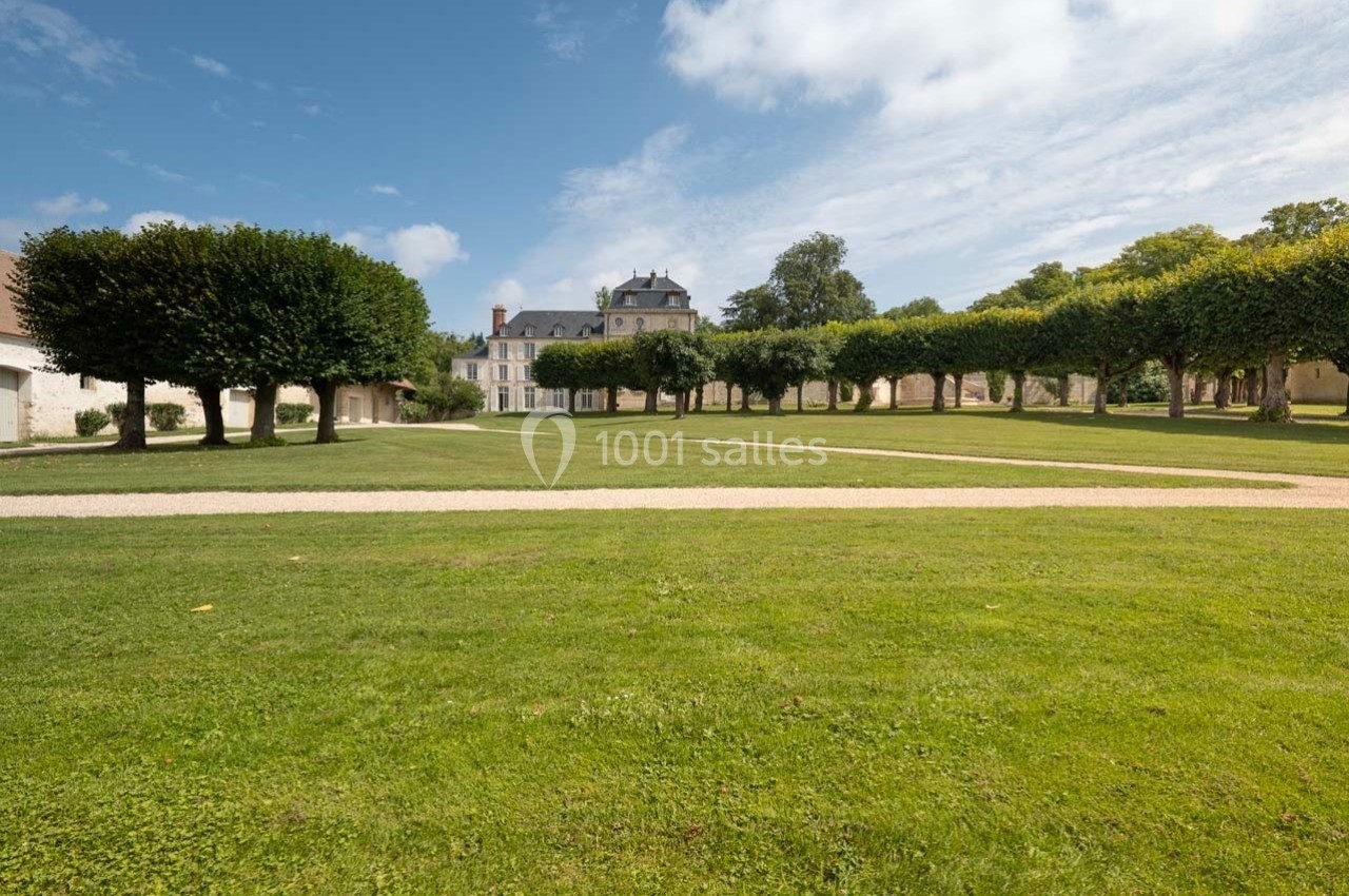 Jardin avec pelouse bien entretenue, allées gravillonnées et rangées d'arbres, devant un bâtiment ancien sous un ciel dégagé.