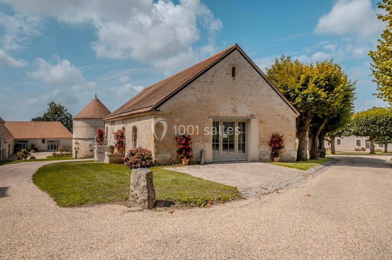 Bâtiment en pierre avec toit en pente, entouré d'arbres, de fleurs et d'une allée gravillonnée sous un ciel partiellement…