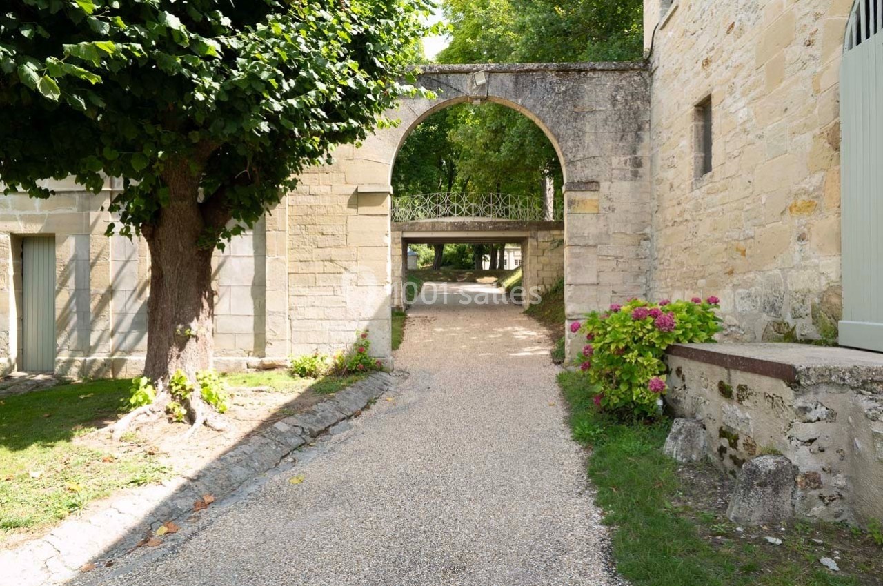 Allée gravillonnée bordée de verdure menant sous une arche en pierre, avec des bâtiments anciens en arrière-plan.