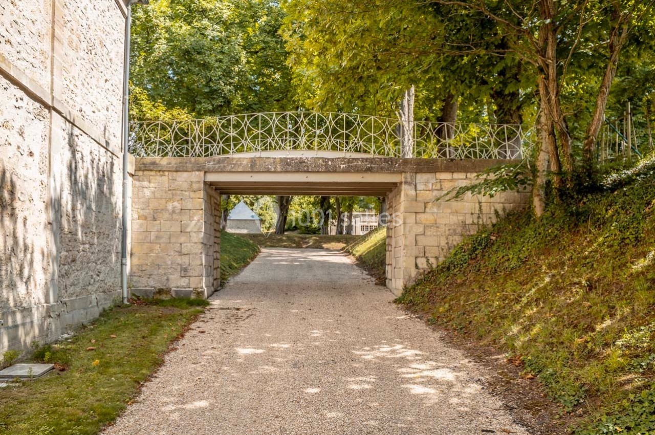 Pont en pierre avec garde-corps en fer forgé surplombant un chemin gravillonné entouré de verdure.