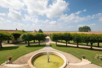 Grand bâtiment classique entouré d'une pelouse bien entretenue, avec un arbre imposant et un petit bassin au premier plan.