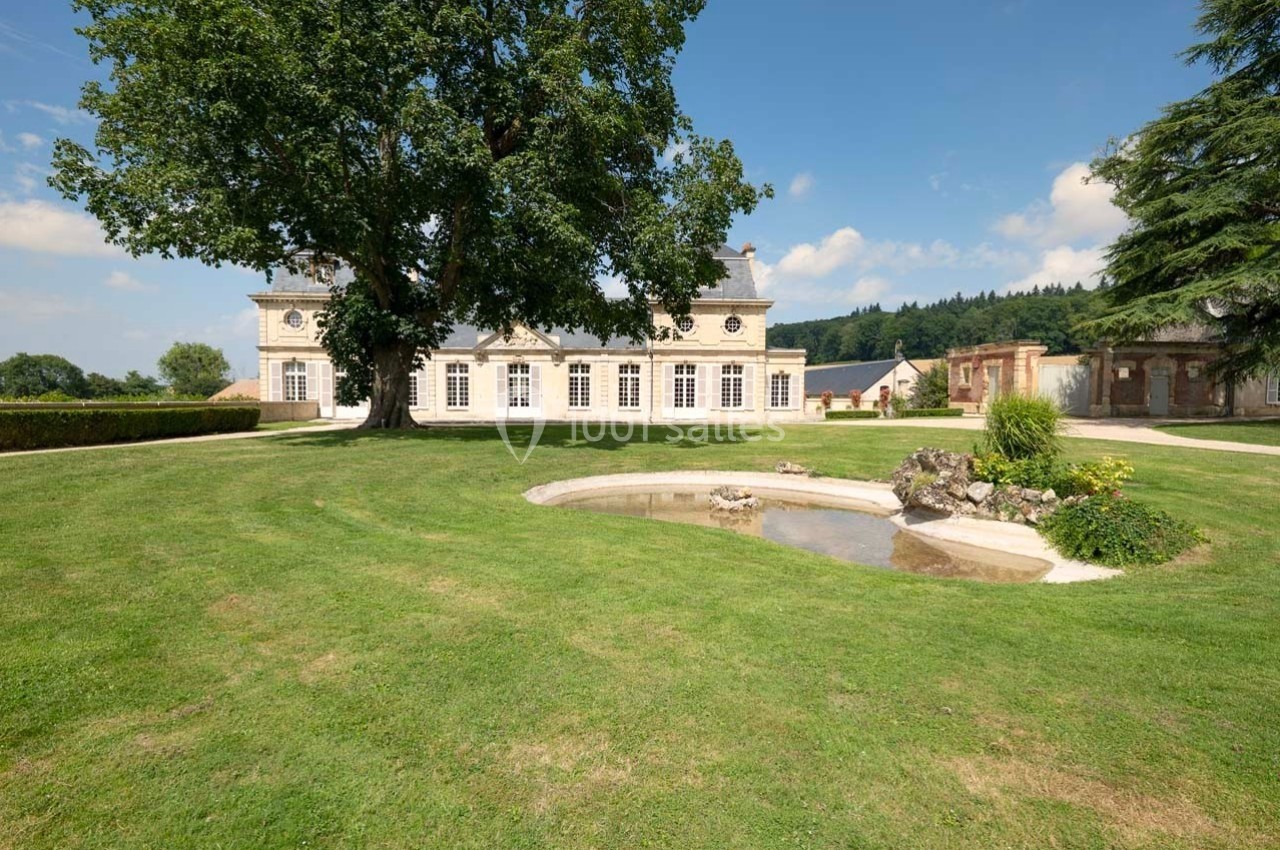 Grand bâtiment classique entouré d'une pelouse bien entretenue, avec un arbre imposant et un petit bassin au premier plan.