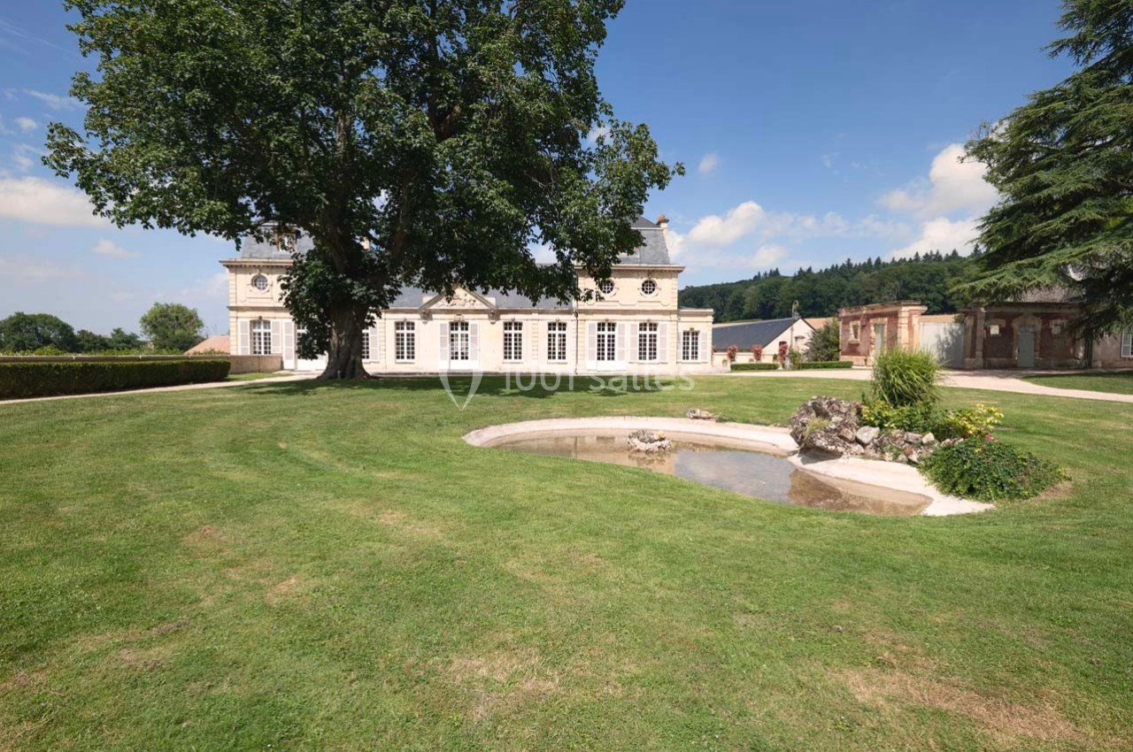 Manoir entouré d'un grand jardin verdoyant avec un arbre imposant et un petit bassin au premier plan.