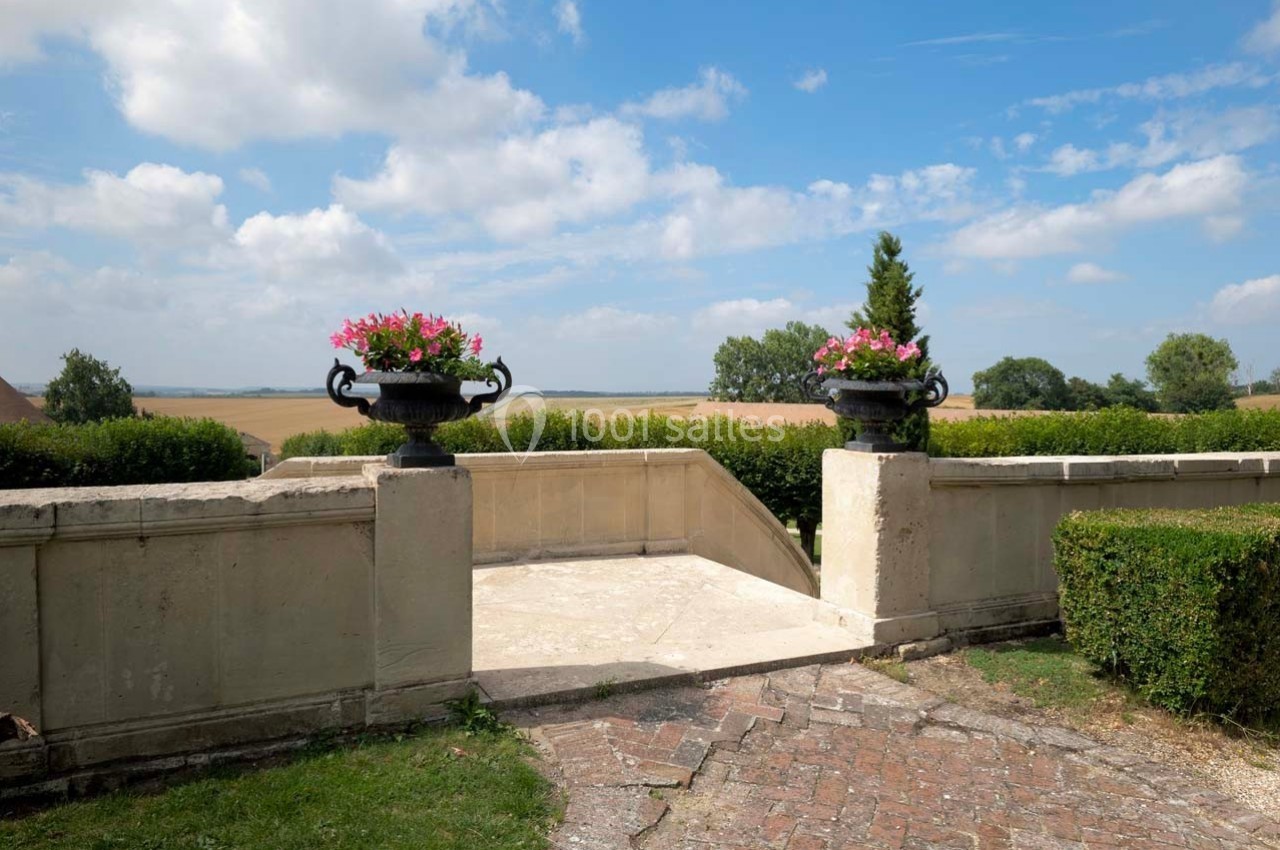Vue d'une terrasse en pierre avec des jardinières fleuries, donnant sur un paysage rural sous un ciel partiellement nuageux.