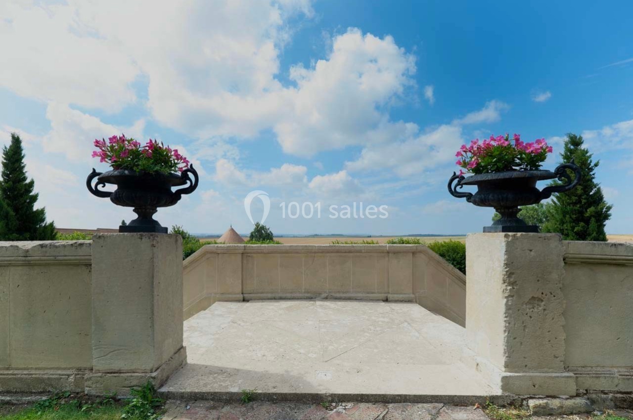 Deux jardinières fleuries sur des piliers encadrent une terrasse offrant une vue dégagée sur la campagne sous un ciel bleu.