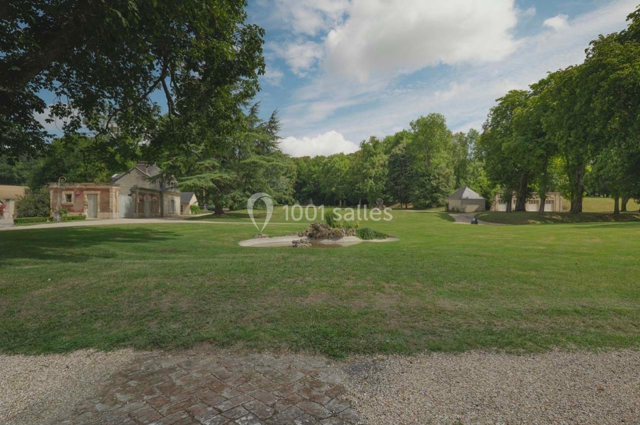 Grand jardin verdoyant avec pelouse, arbres, petite fontaine centrale et bâtiments en pierre en arrière-plan.
