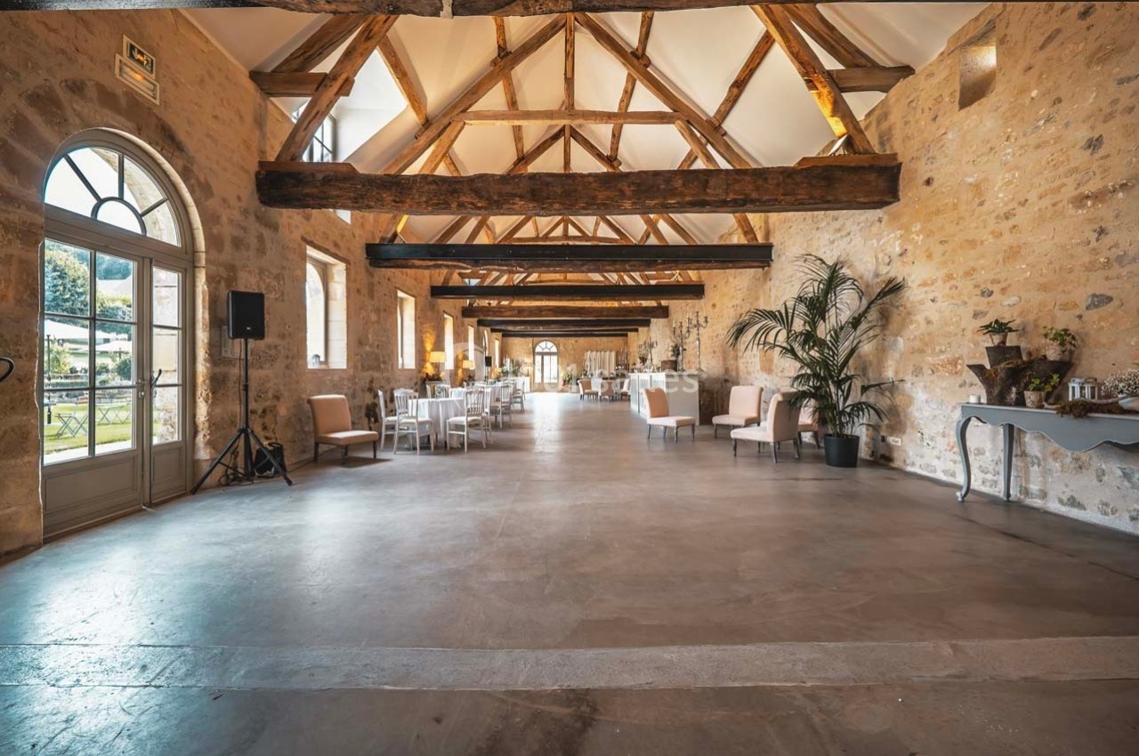 Salle lumineuse avec poutres apparentes, murs en pierre, grandes fenêtres et mobilier disposé pour un événement.