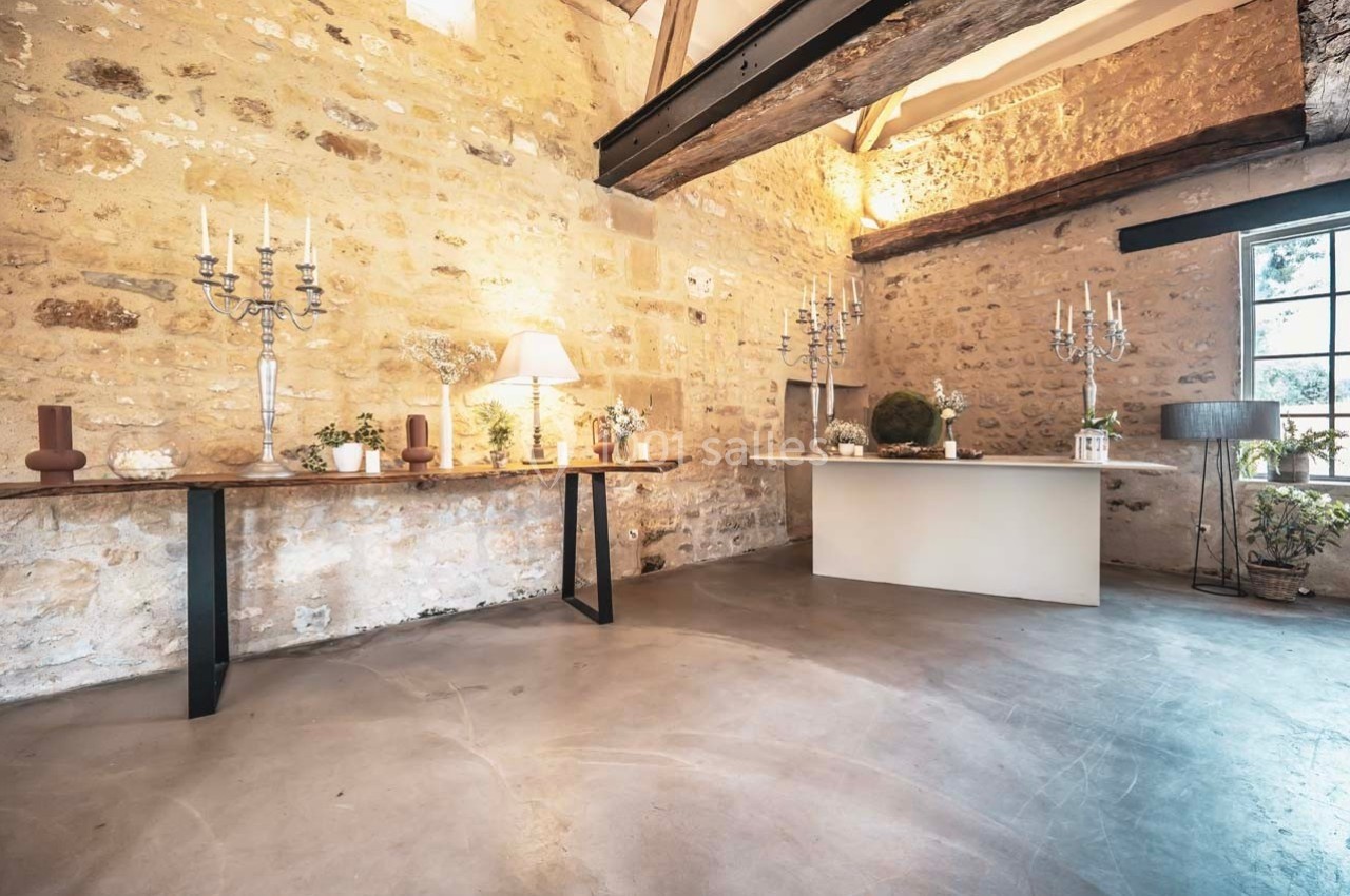 Salle en pierre avec poutres apparentes, tables décorées de chandeliers et plantes, éclairée par une fenêtre.