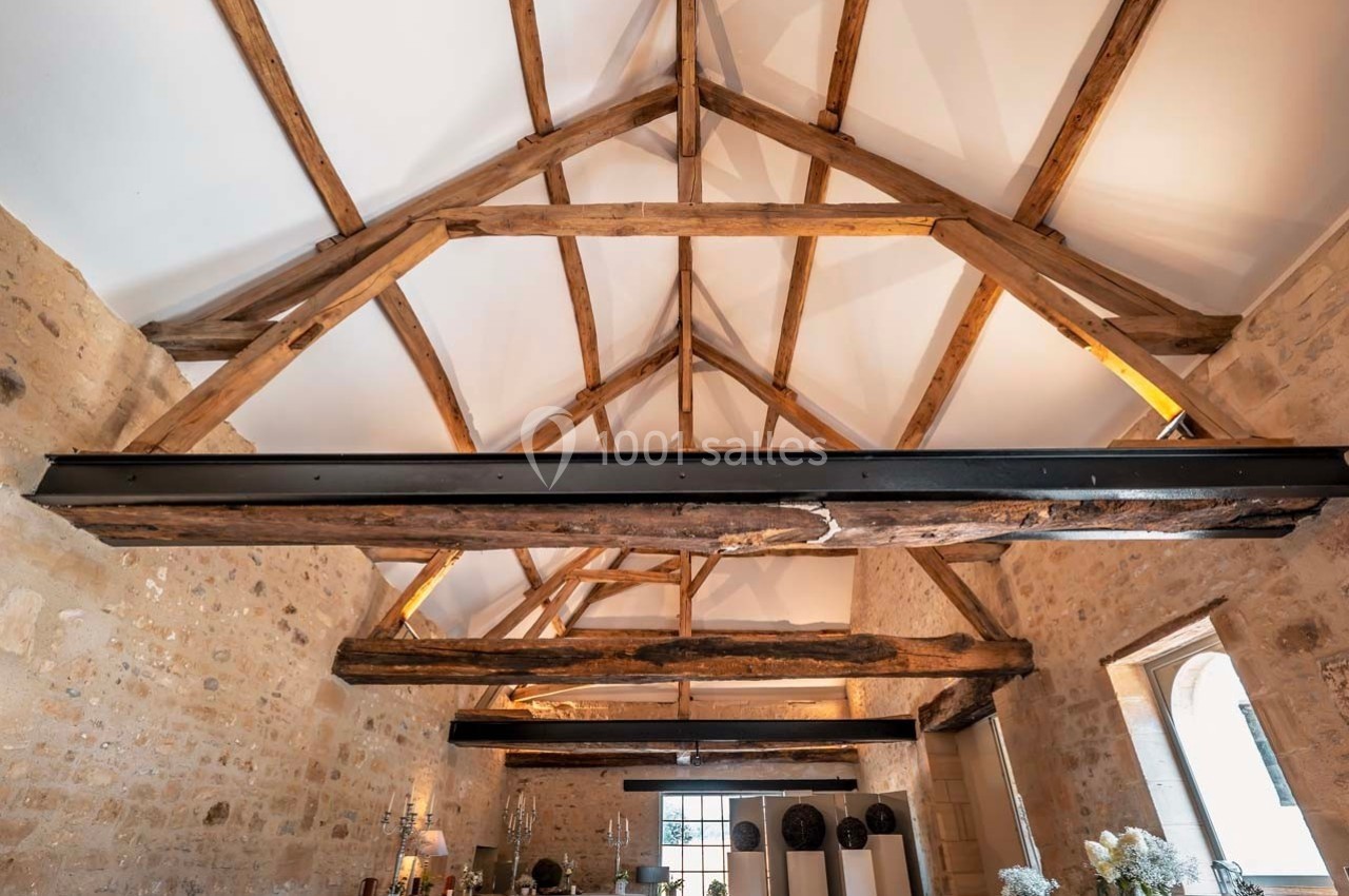 Plafond avec poutres apparentes en bois dans une pièce en pierre, éclairée par des fenêtres et des lumières naturelles.