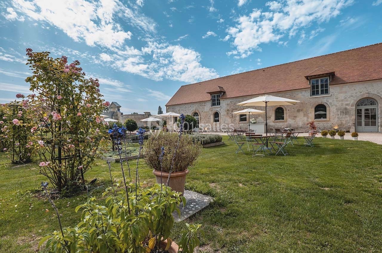 Jardin avec tables, chaises et parasols devant un bâtiment en pierre sous un ciel partiellement nuageux.