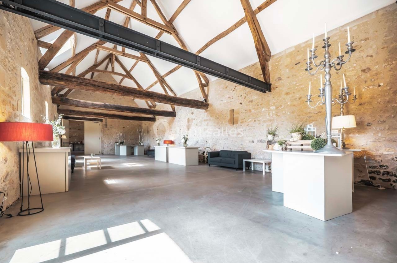 Salle lumineuse avec murs en pierre, poutres apparentes, mobilier moderne et grandes fenêtres laissant entrer la lumière.