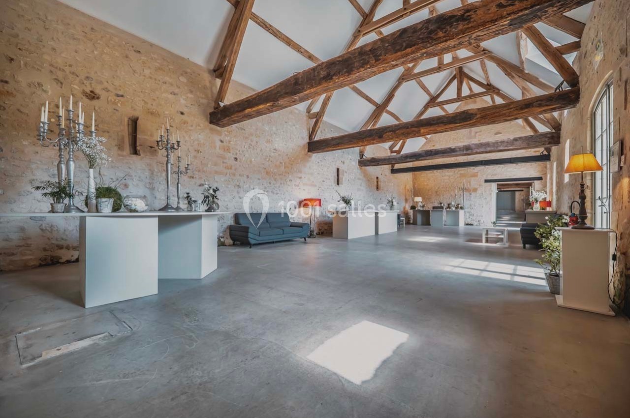 Salle spacieuse avec murs en pierre, poutres apparentes, mobilier moderne et lumière naturelle entrant par les fenêtres.