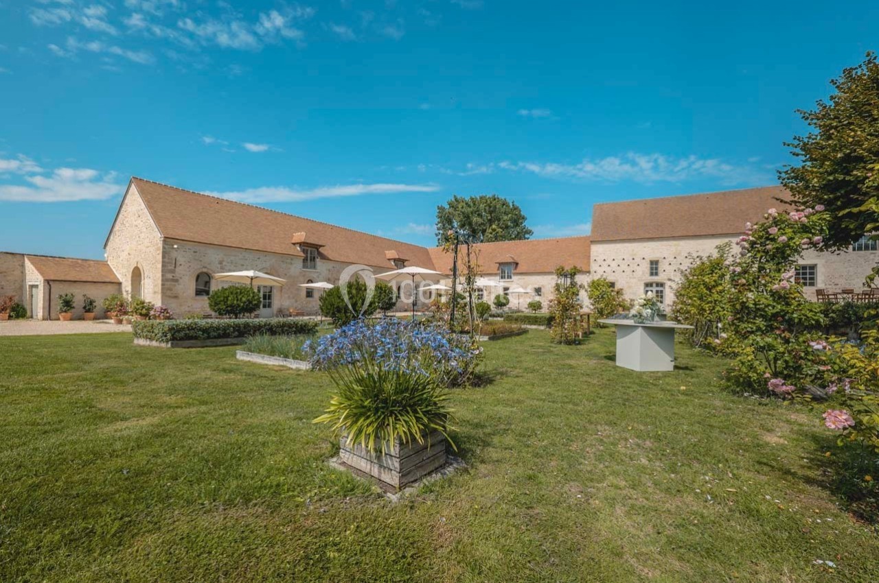 Bâtiments en pierre avec toits en tuiles entourés d'un jardin fleuri et d'une pelouse sous un ciel bleu.