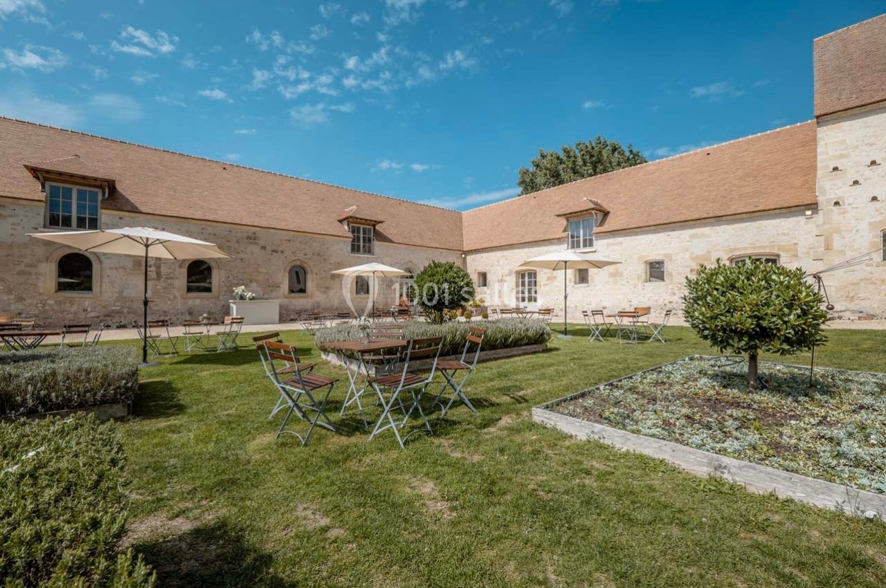 Cour intérieure avec tables, chaises en métal, parasols, pelouse et parterres de fleurs, entourée de bâtiments en pierre.
