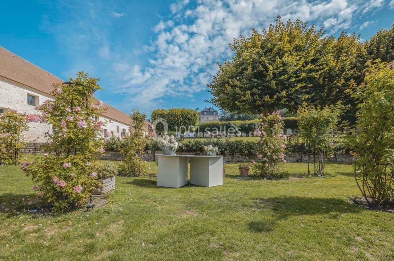 Jardin fleuri avec des rosiers, une table blanche et un bâtiment en pierre sous un ciel bleu.
