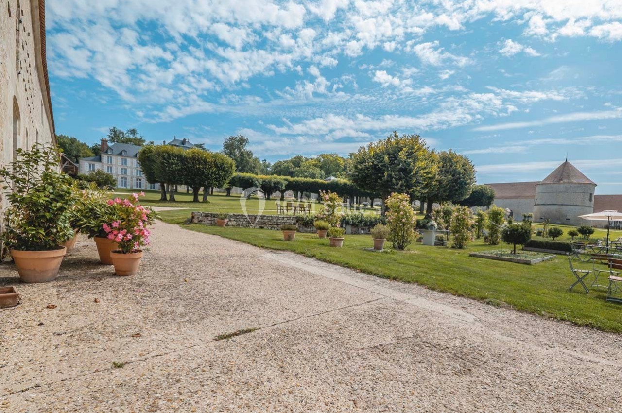 Allée en gravier bordée de pots de fleurs, menant à un jardin avec pelouse, arbres et bâtiments en arrière-plan.