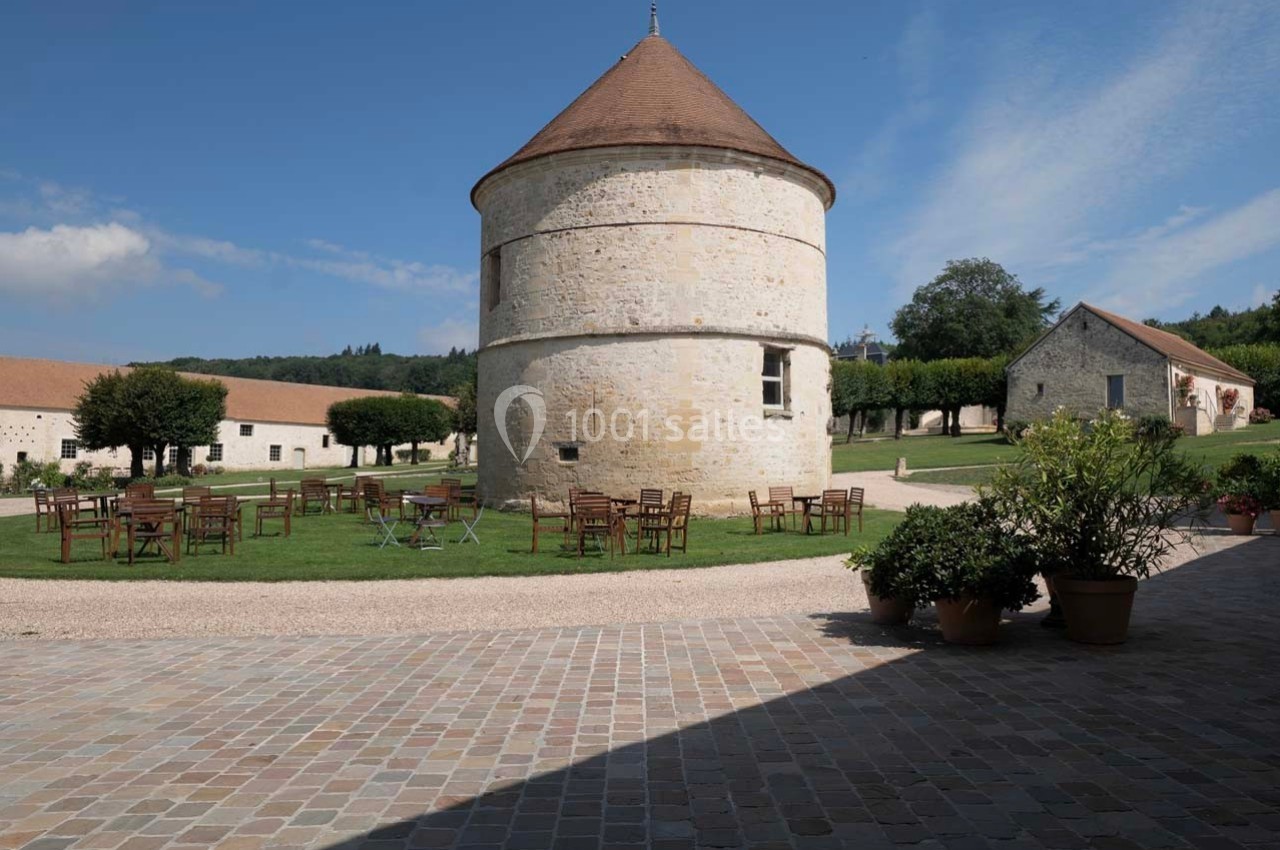 Tour en pierre ronde avec toit conique, entourée de tables en plein air sur une pelouse, dans un cadre rural.
