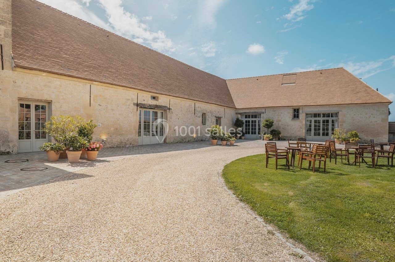 Cour extérieure d'une bâtisse en pierre avec toit en tuiles, tables et chaises sur une pelouse, par temps ensoleillé.