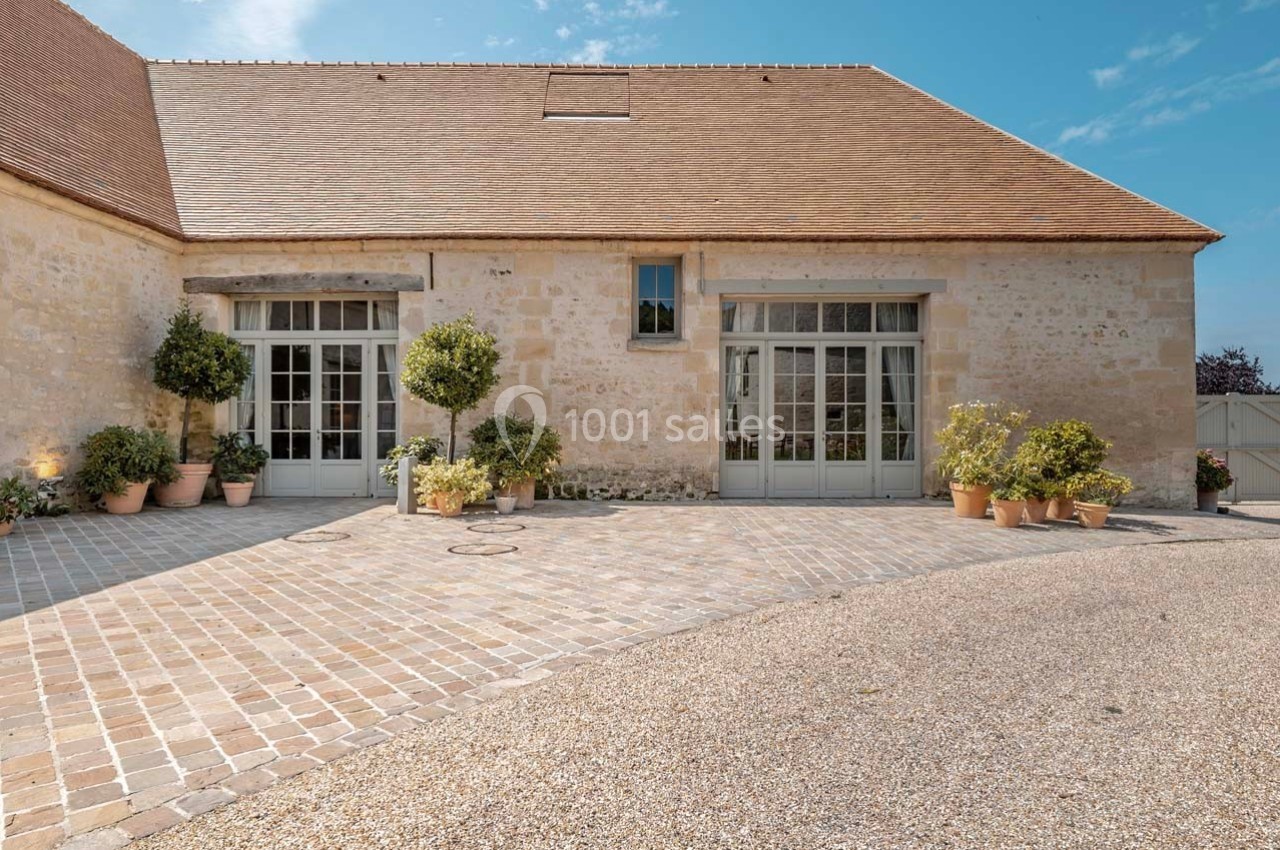 Façade d'un bâtiment en pierre avec grandes portes vitrées, entouré de plantes en pots sur une cour pavée.