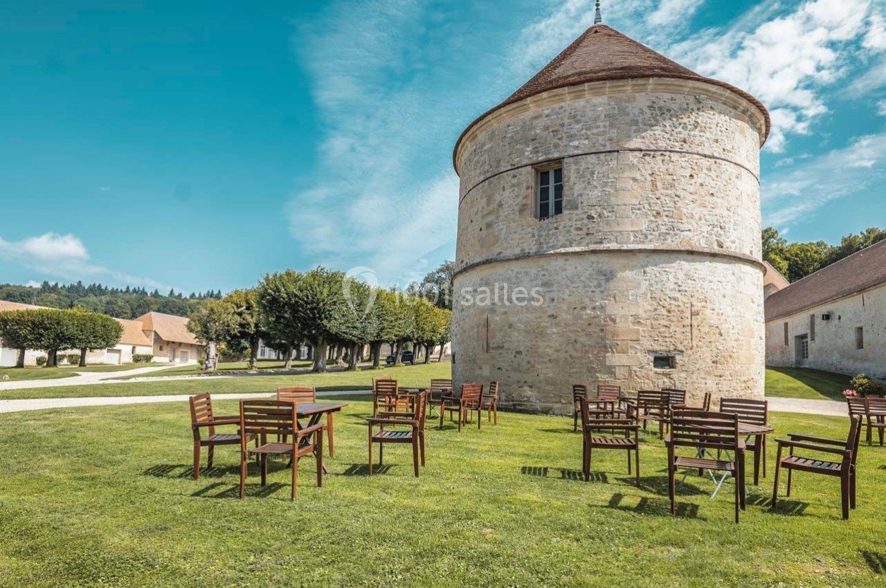 Chaises et tables en bois disposées sur une pelouse devant une tour en pierre dans un cadre rural ensoleillé.