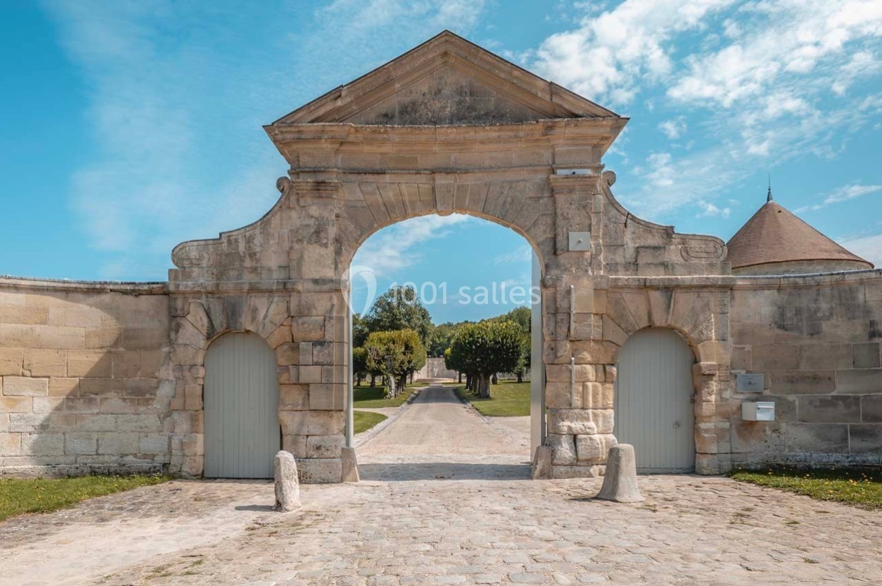 Porte en pierre ancienne avec une arche centrale, donnant sur une allée bordée d'arbres sous un ciel bleu.