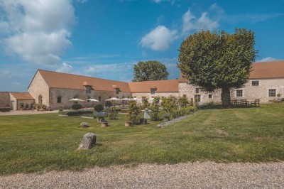 Petit pont en pierre surplombant un chemin, entouré de verdure et jouxtant un bâtiment ancien avec toiture en ardoise.